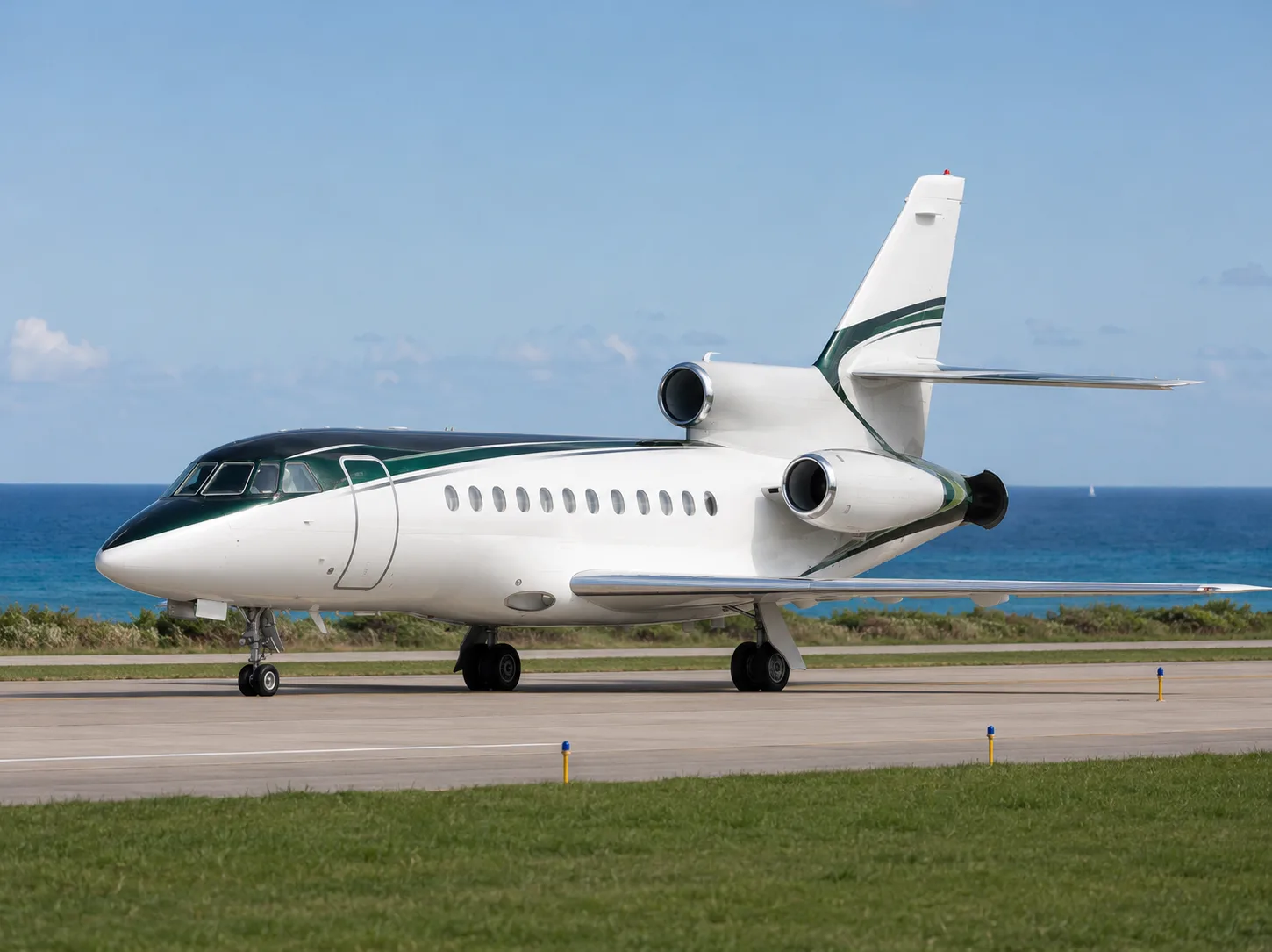 A white and dark green Dassault Falcon business jet is parked on a runway with the ocean in the background.