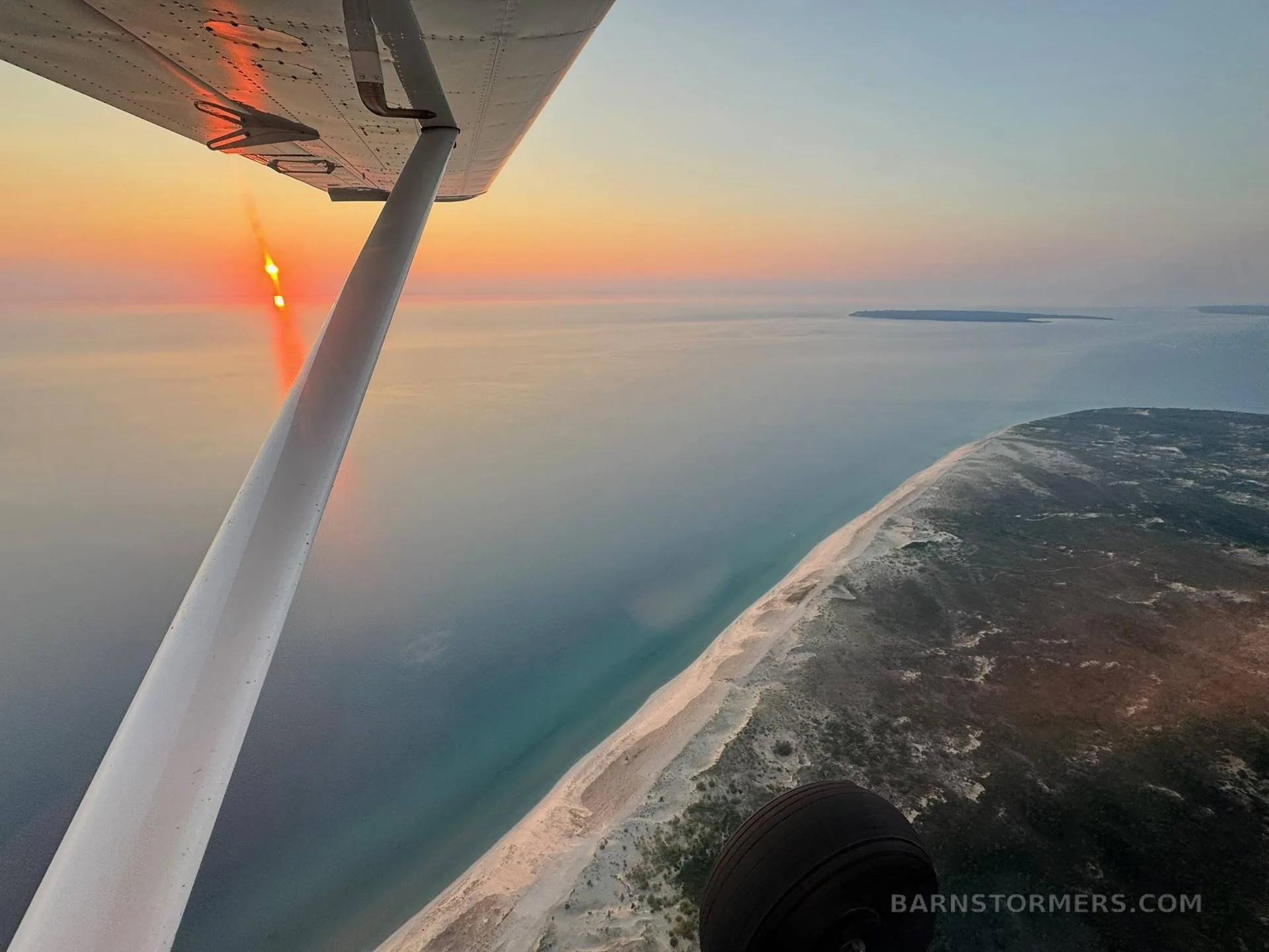 An aerial view from a small aircraft showing its wing and wheel over a sandy coastline and ocean at sunset.