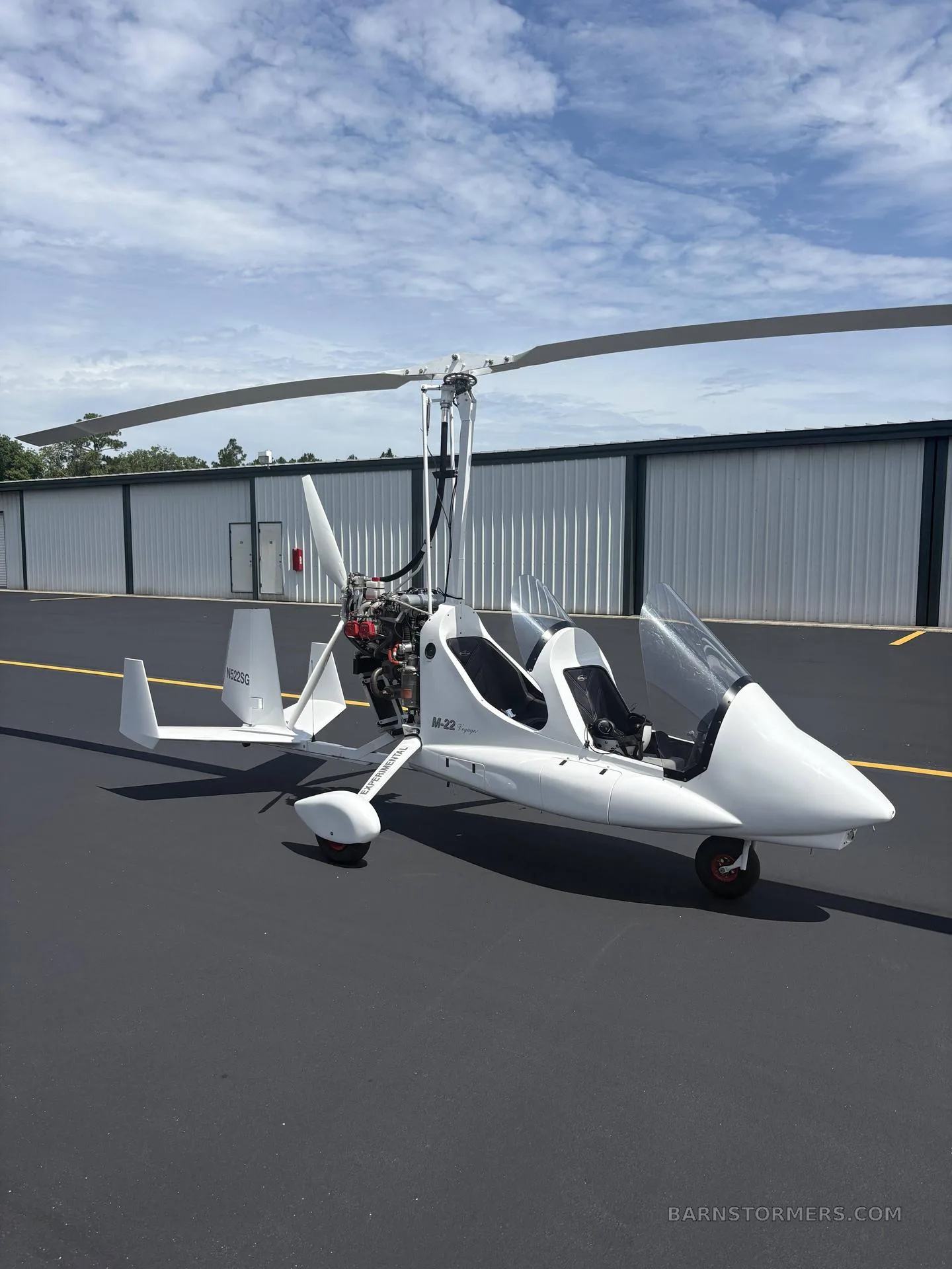 A white M-22 gyrocopter with an open cockpit is parked on an asphalt tarmac in front of a hangar under a partly cloudy sky.