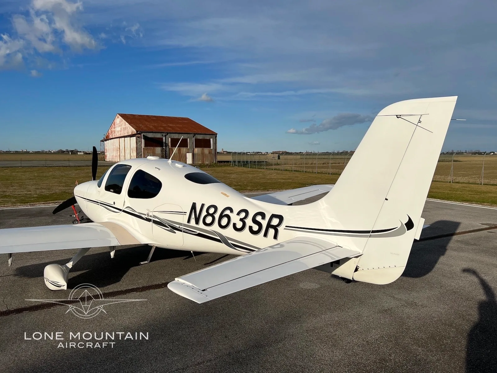 A white Cirrus SR22 aircraft with registration N863SR is parked on an airfield, with a red-roofed building in the background.