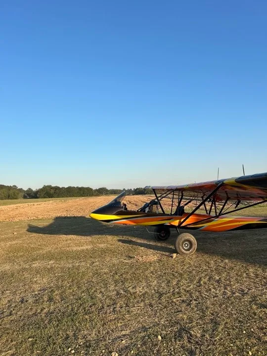 A side view of an orange and black ultralight aircraft with large tires, parked in a dry grassy field under a clear blue sky.