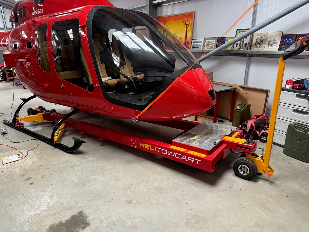 The front of a red Bell 505 helicopter is shown inside a hangar, mounted on a red HELITOWCART.