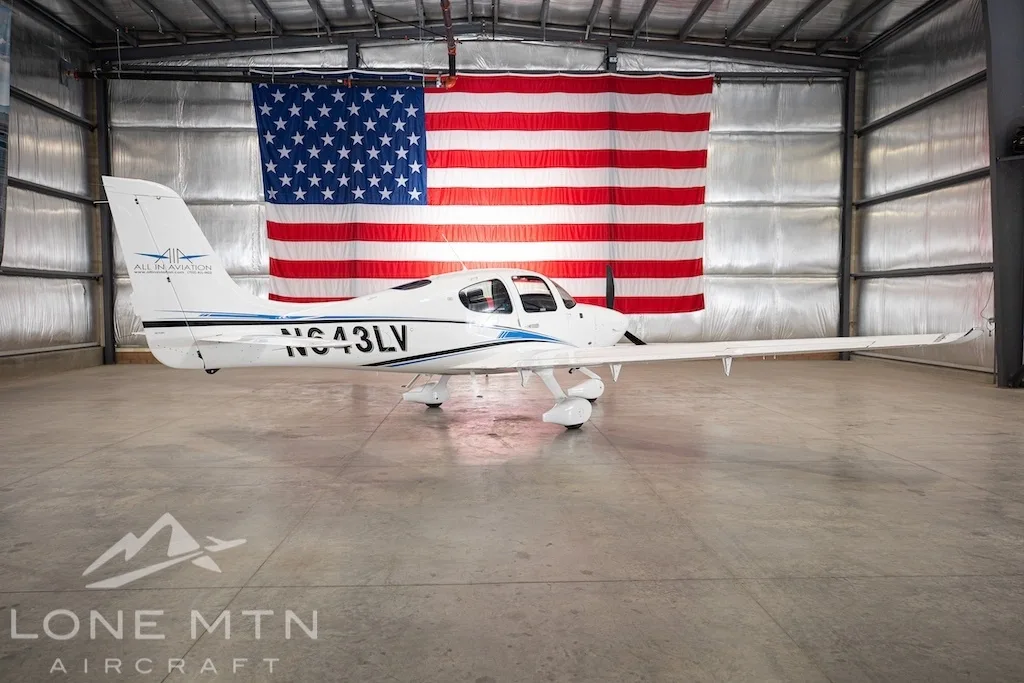 A white Cirrus SR22 aircraft with blue and black stripes, tail number N643LV, parked in a hangar with an American flag in the background.