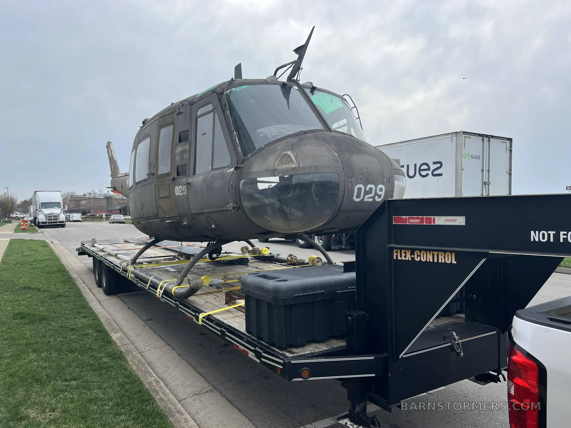 A dark green Bell UH-1 Iroquois helicopter, with "029" on the nose, loaded onto a flatbed trailer on a cloudy day.