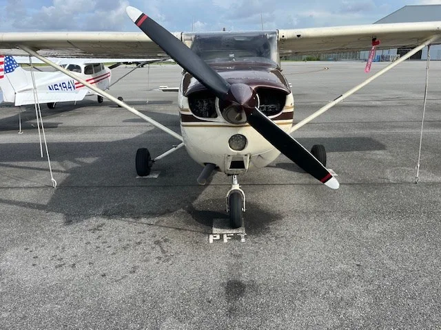 Front view of a brown and white Cessna aircraft with a propeller, parked on an asphalt tarmac.