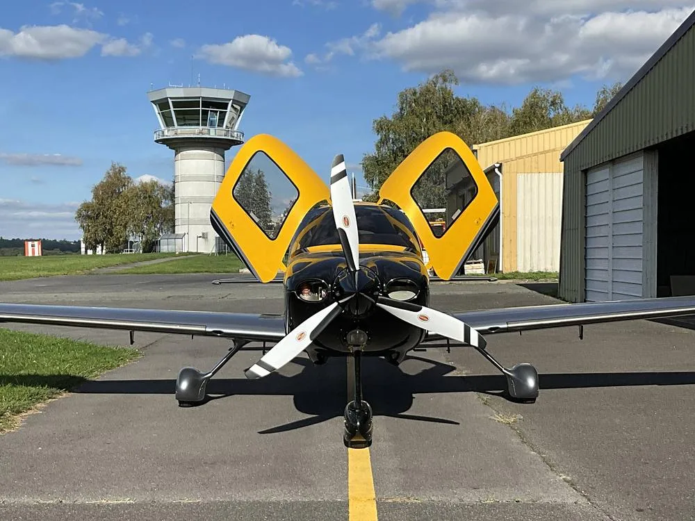 Front view of a yellow and black Cirrus SR22 aircraft with its gull-wing doors open, parked in front of an air traffic control tower.