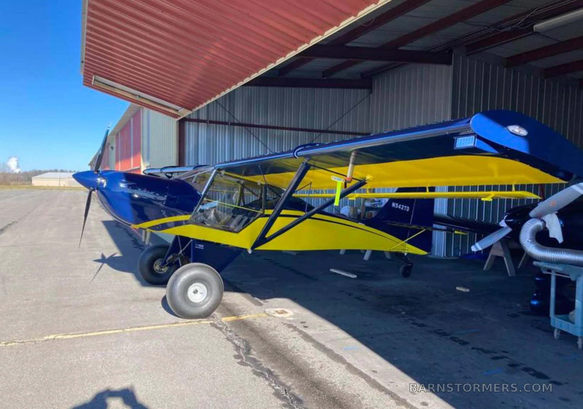 A blue and yellow high-wing aircraft with tundra tires parked partially inside a hangar on a sunny day.