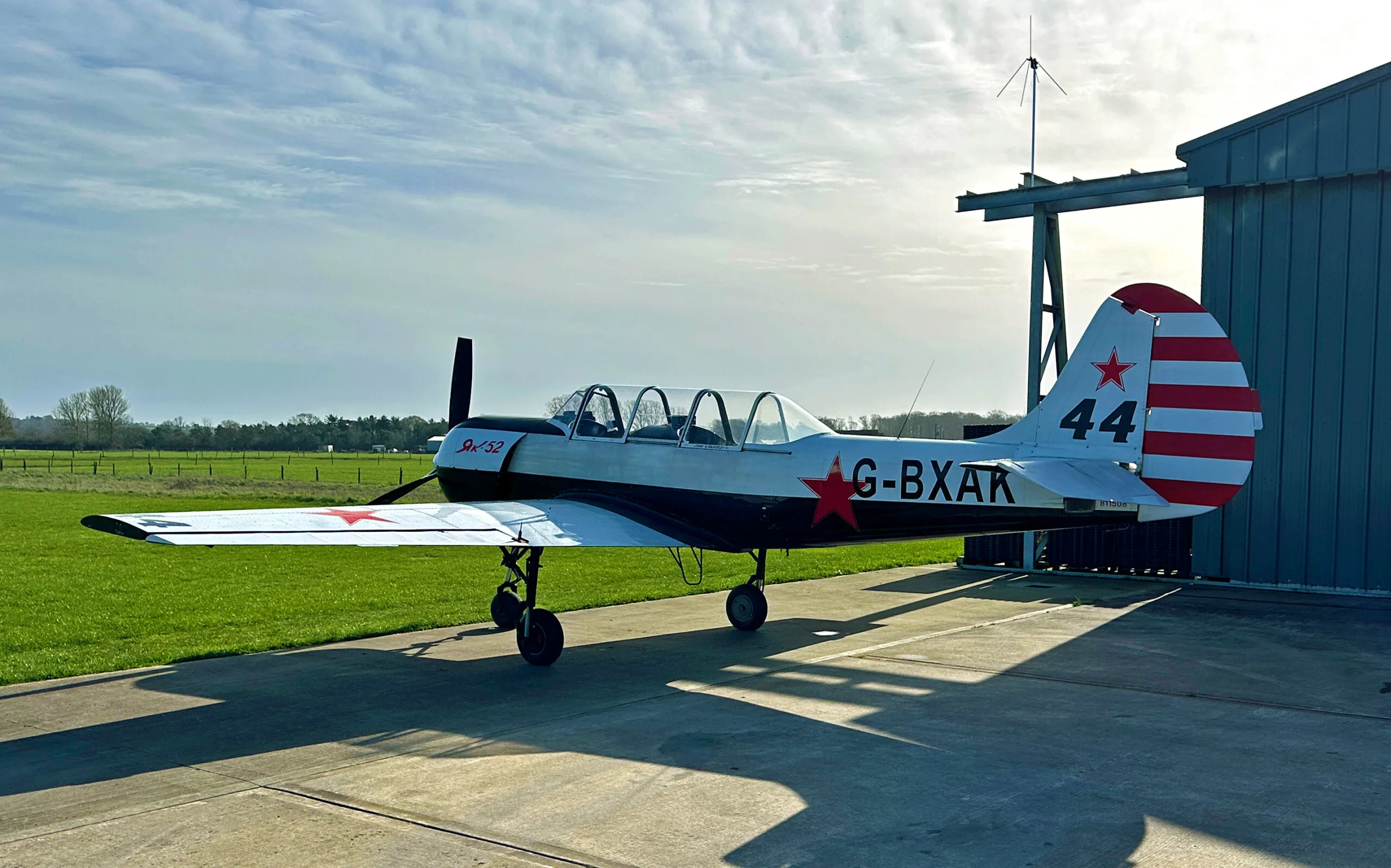 A white and black Yak-52 aircraft with red stars and tail stripes, parked on a concrete pad next to a hangar and a grassy field.