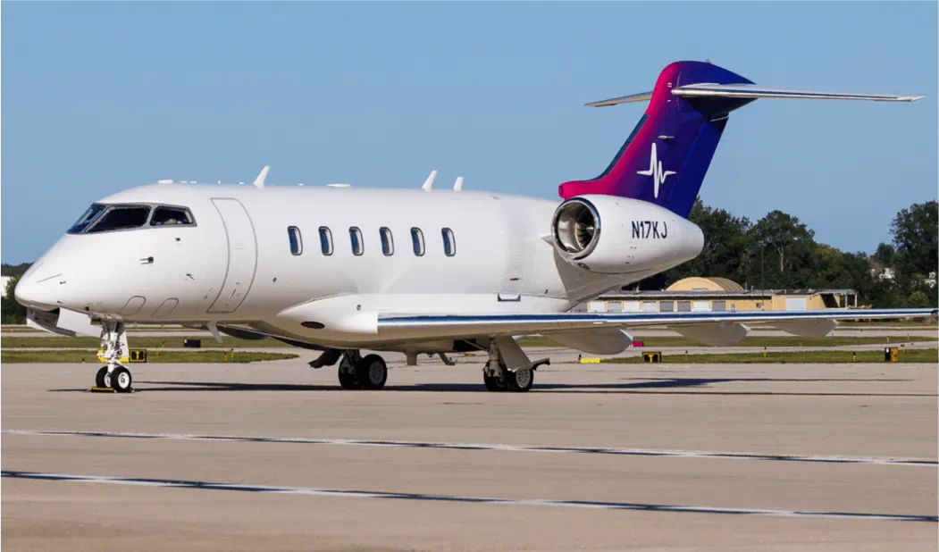 A white Bombardier Challenger 350 business jet with a purple and blue tail livery featuring a heartbeat symbol, parked on an airfield.