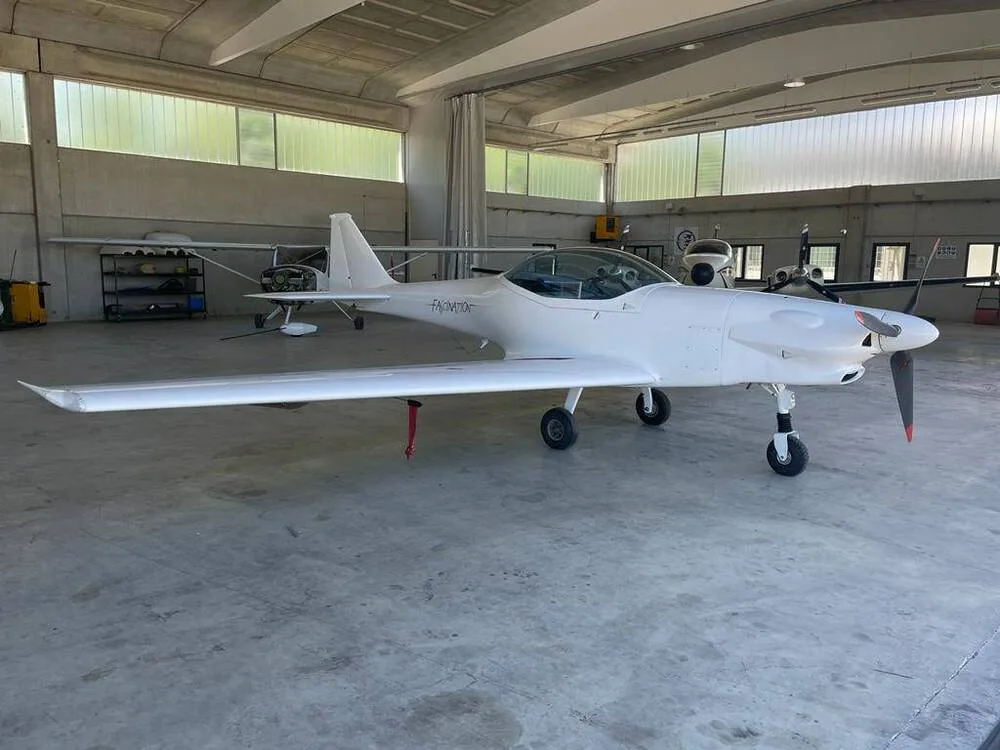 A white Fascination ultralight aircraft with a clear canopy is parked inside a hangar, viewed from the front-right.