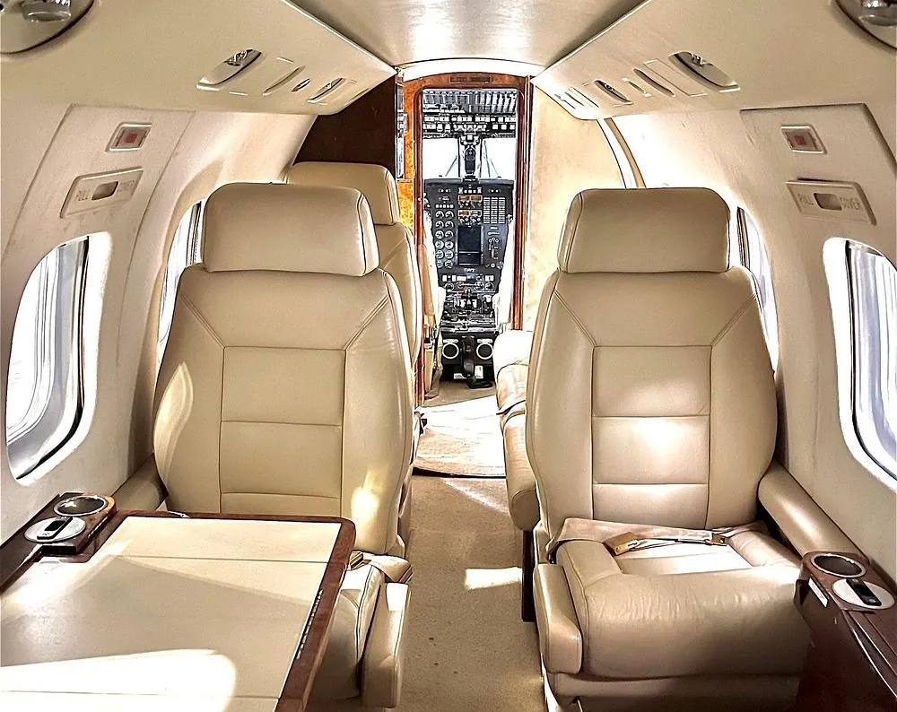 Interior view of a private jet cabin with beige leather seats, looking forward towards the cockpit.