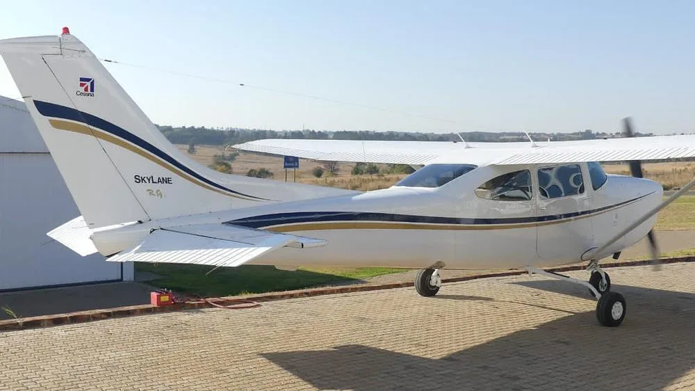A white Cessna Skylane aircraft with blue and gold stripes and "Skylane R.G." text on the tail, parked on a paved surface outdoors.