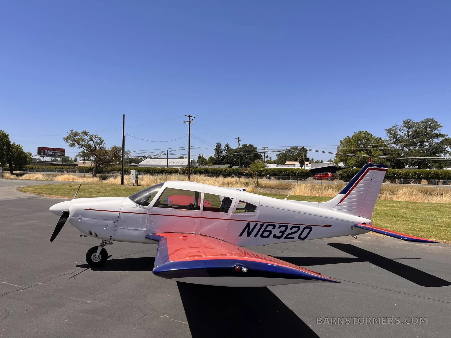 A white Piper Cherokee aircraft with red and blue accents on the wings and tail, parked on an asphalt ramp under a clear blue sky.