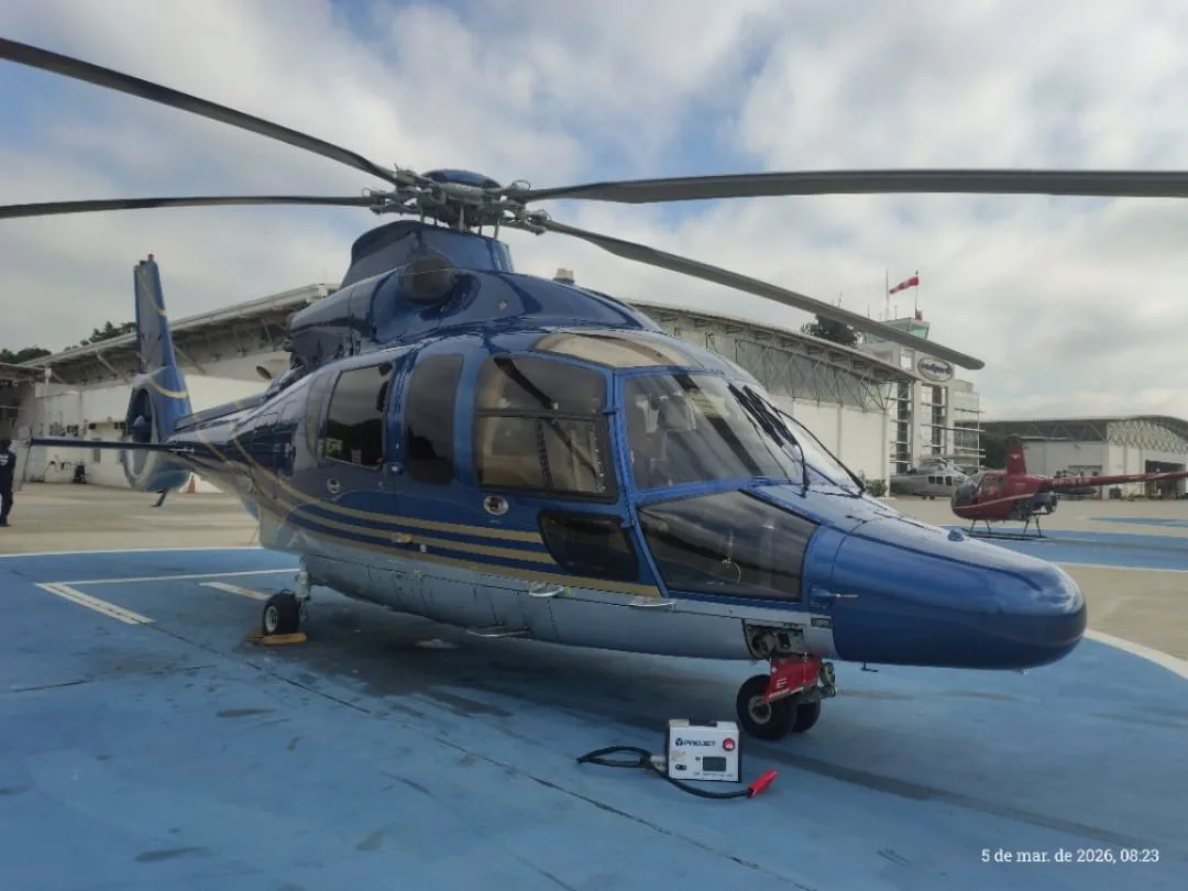 A blue and silver helicopter with gold stripes is parked on a blue helipad, viewed from the front-left, with a building in the background.