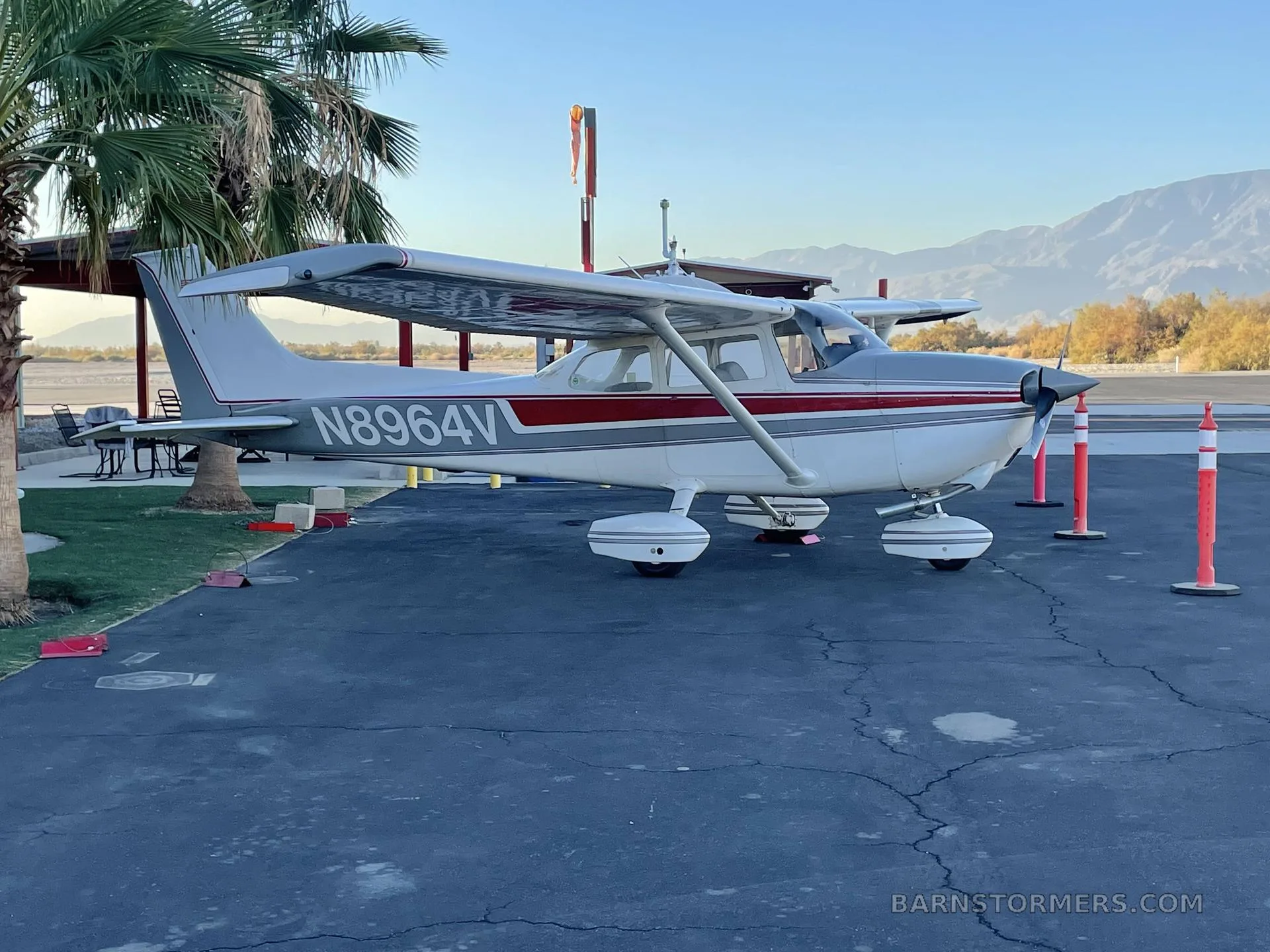 A white and grey Cessna 172M with red stripes, registration N8964V, parked on an airfield with palm trees and mountains in the background.