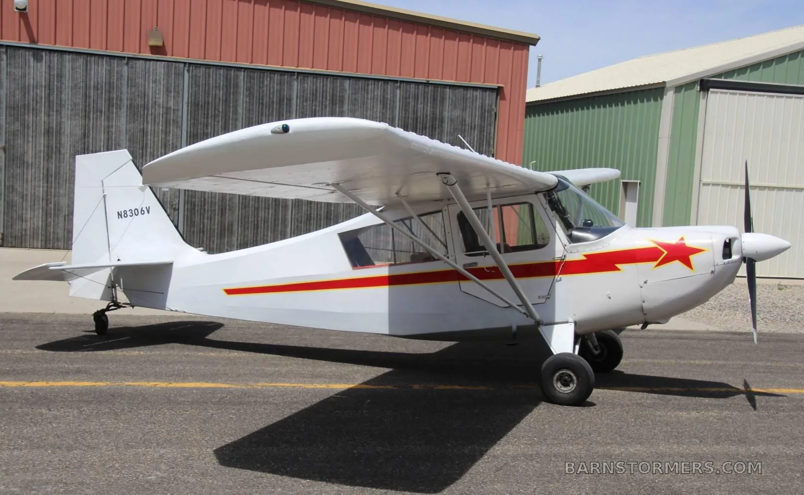 A white single-engine high-wing aircraft with red stripes and a red star on the cowl, parked on an asphalt ramp next to hangars.