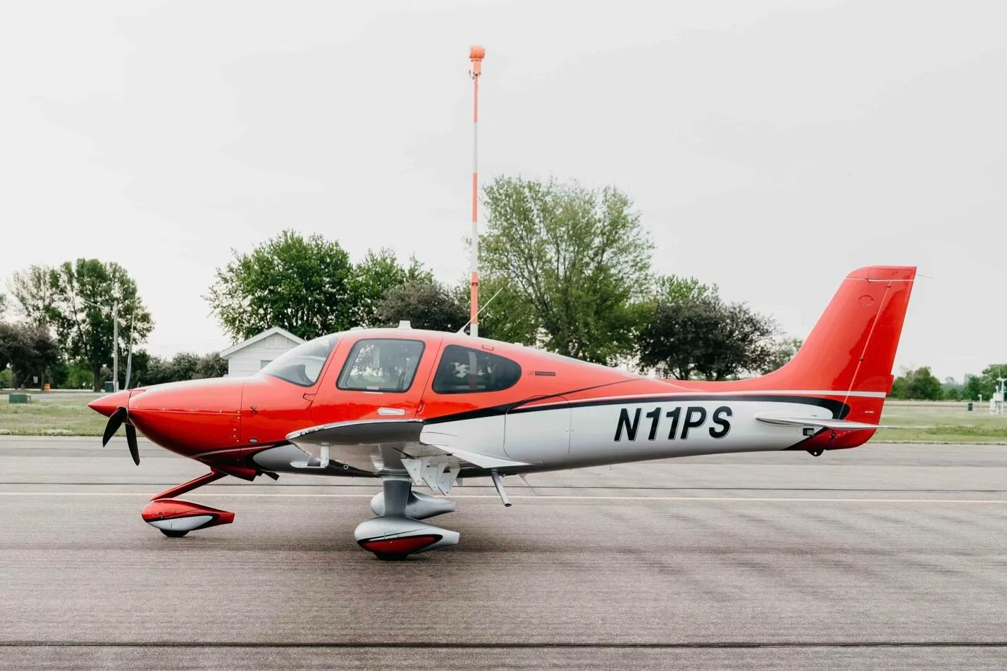 A red and silver Cirrus SR20 Premium aircraft (N11PS) parked on an airfield tarmac with a beacon and trees in the background.