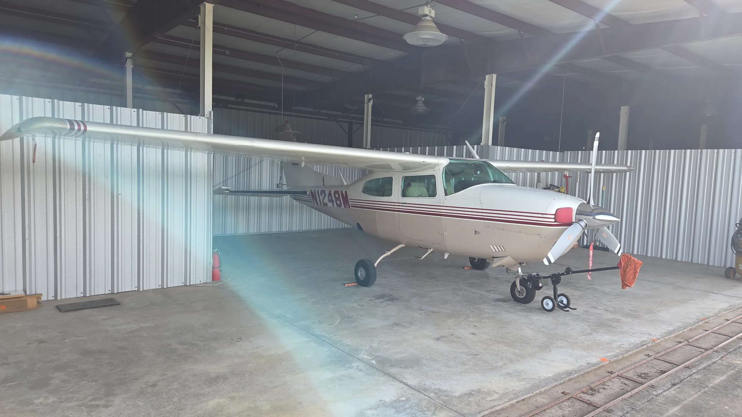 A side view of a white and tan single-engine propeller aircraft with red stripes and registration N1248M, parked in a hangar with lens flare.