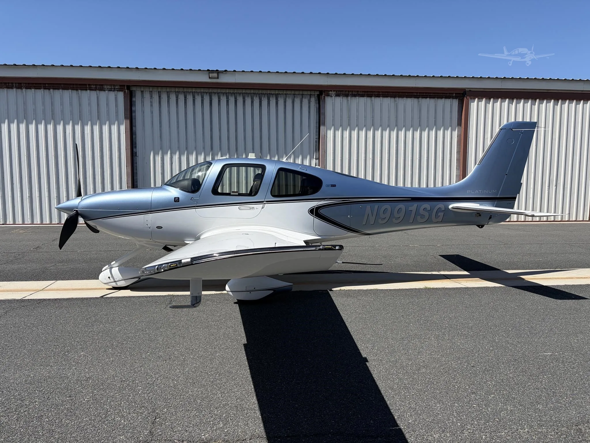 A blue and white Cirrus SR22 aircraft, registration N991SG, is parked on an asphalt tarmac in front of a hangar under a clear sky.