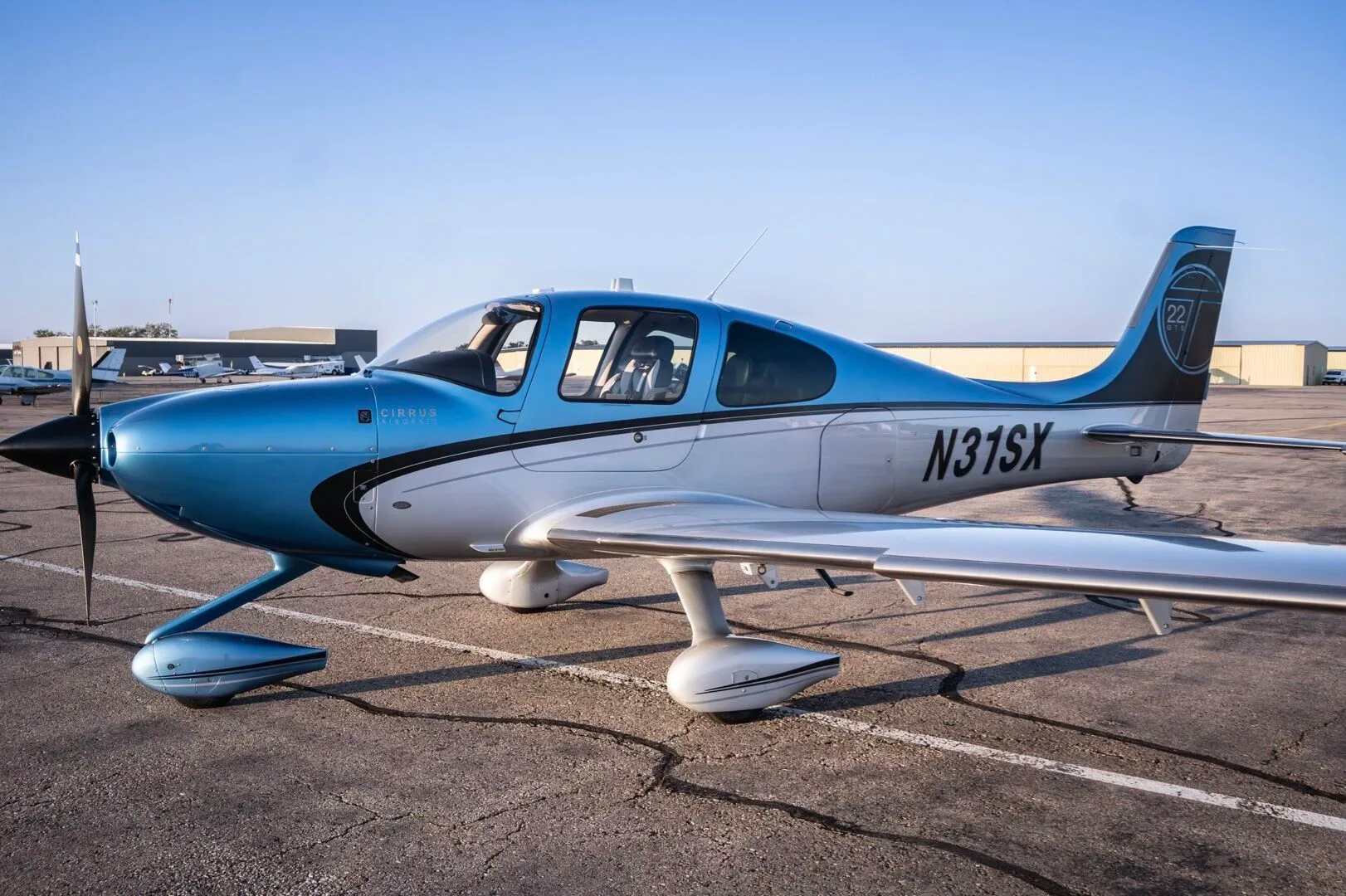 A Cirrus SR22T aircraft, N31SX, in blue and silver paint, parked on an airfield with other aircraft in the background.