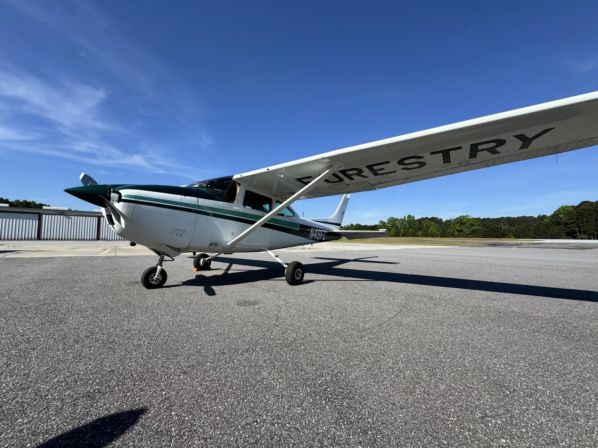 A white and green Cessna 182 Skylane, N49FC, with 'FORESTRY' on its wing, parked on an asphalt apron under a clear blue sky.
