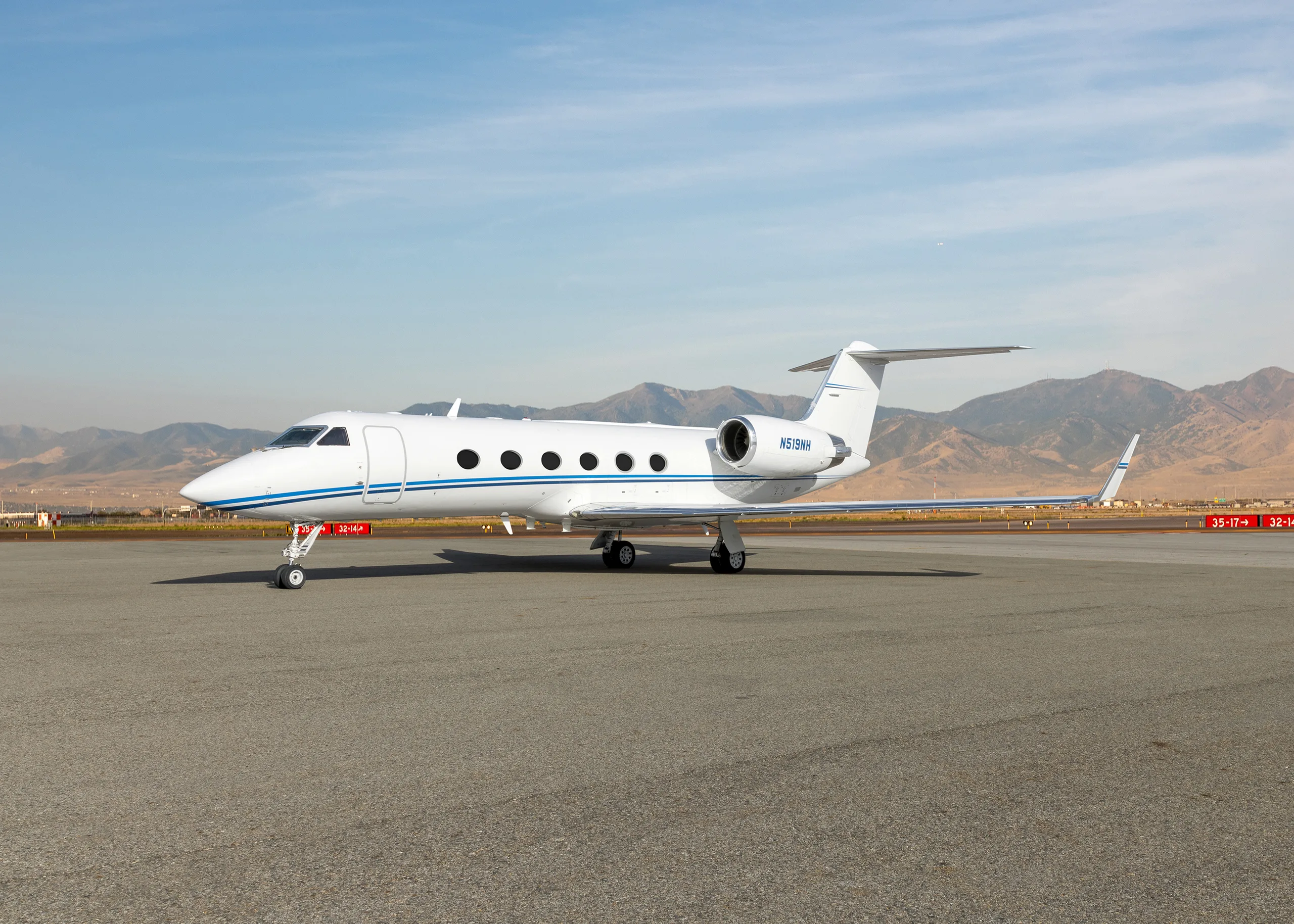 A white Gulfstream G550 business jet with a blue stripe on the tarmac, with mountains in the background.