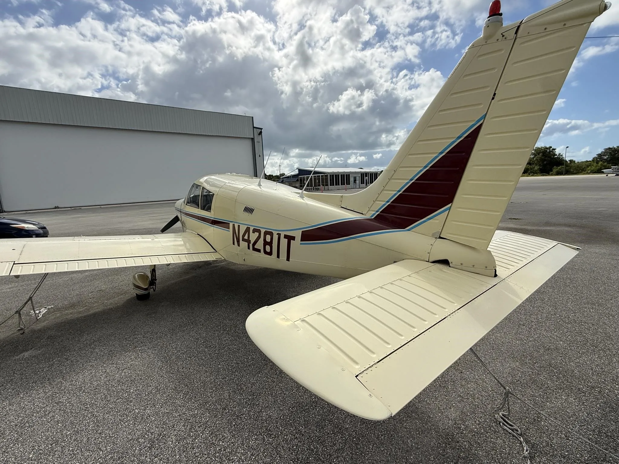 A rear-side view of the cream-colored Piper PA-28-140E Cherokee, registration N4281T, parked on an airfield next to a hangar.