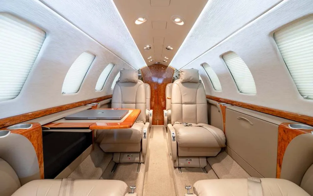 A perspective view of the private jet cabin interior, showing two rows of beige leather seats and a table with a laptop.