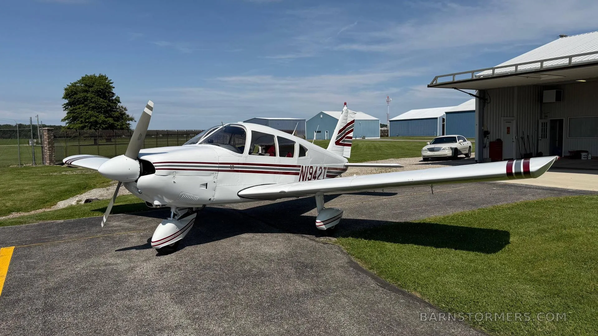 A white single-engine propeller aircraft (N1942T) with red and grey stripes parked on an asphalt ramp in front of hangars on a sunny day.