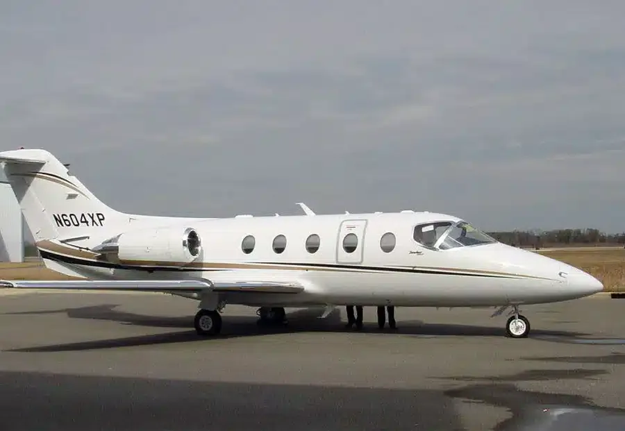 A white Learjet with gold and brown stripes is parked on an asphalt tarmac under a cloudy sky.