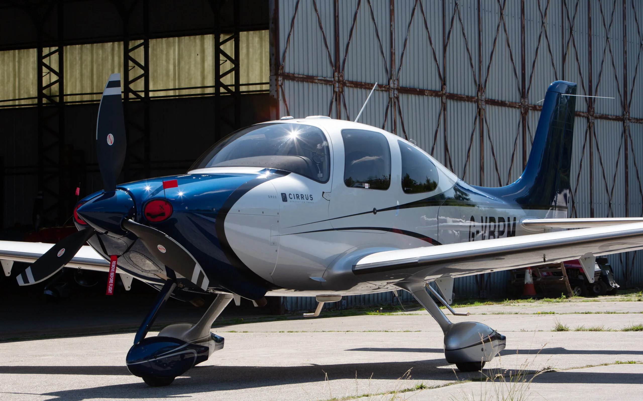 A side view of a blue and white Cirrus SR22 aircraft with registration G-VBPM, parked in front of a hangar.
