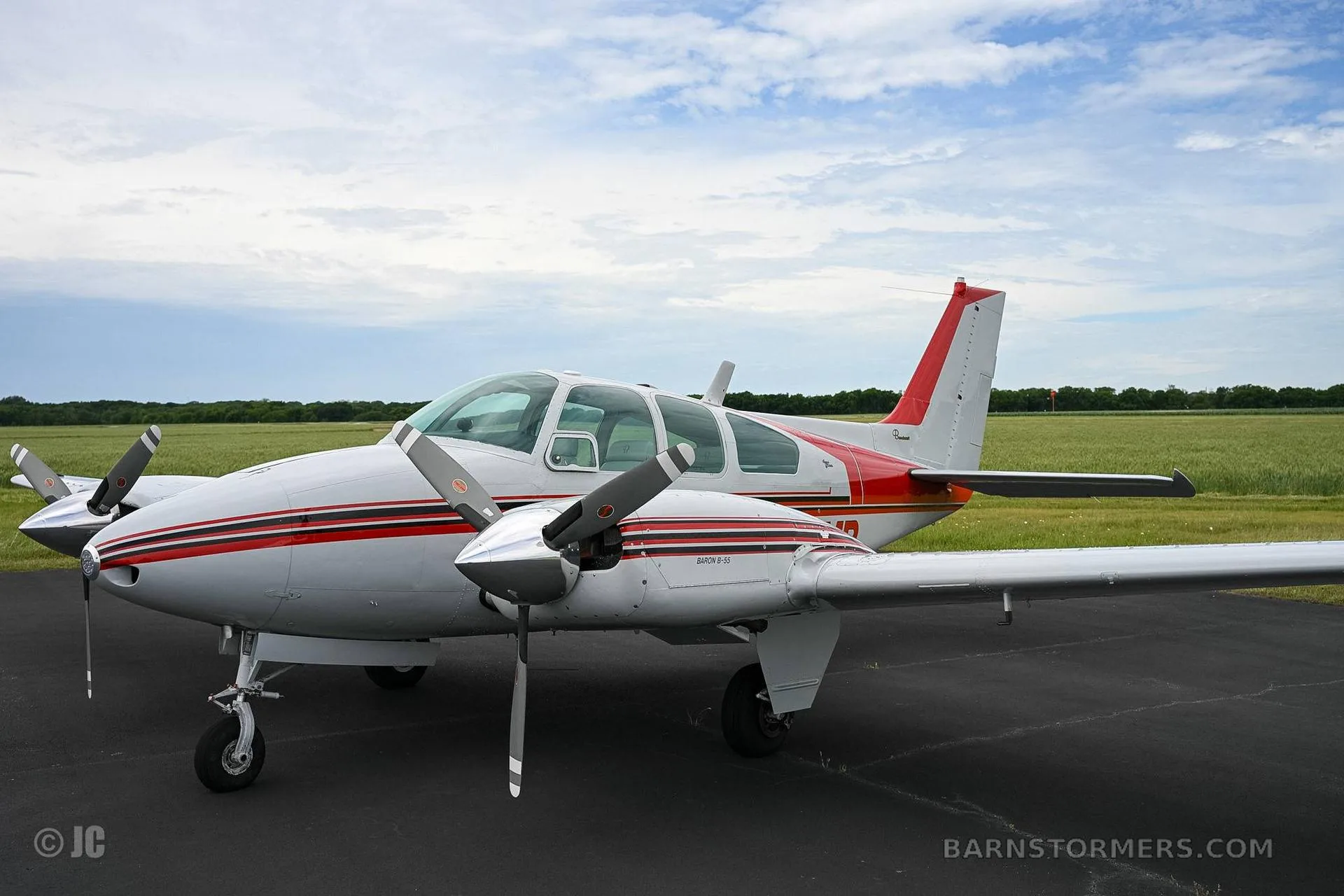 A white and red twin-engine propeller aircraft is parked on an asphalt runway with green fields in the background under a cloudy sky.