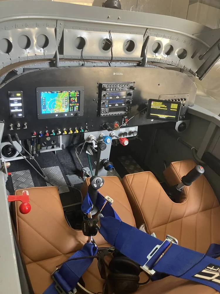 The cockpit of a small aircraft, showing the instrument panel with screens, controls, and two brown leather seats with blue harnesses.