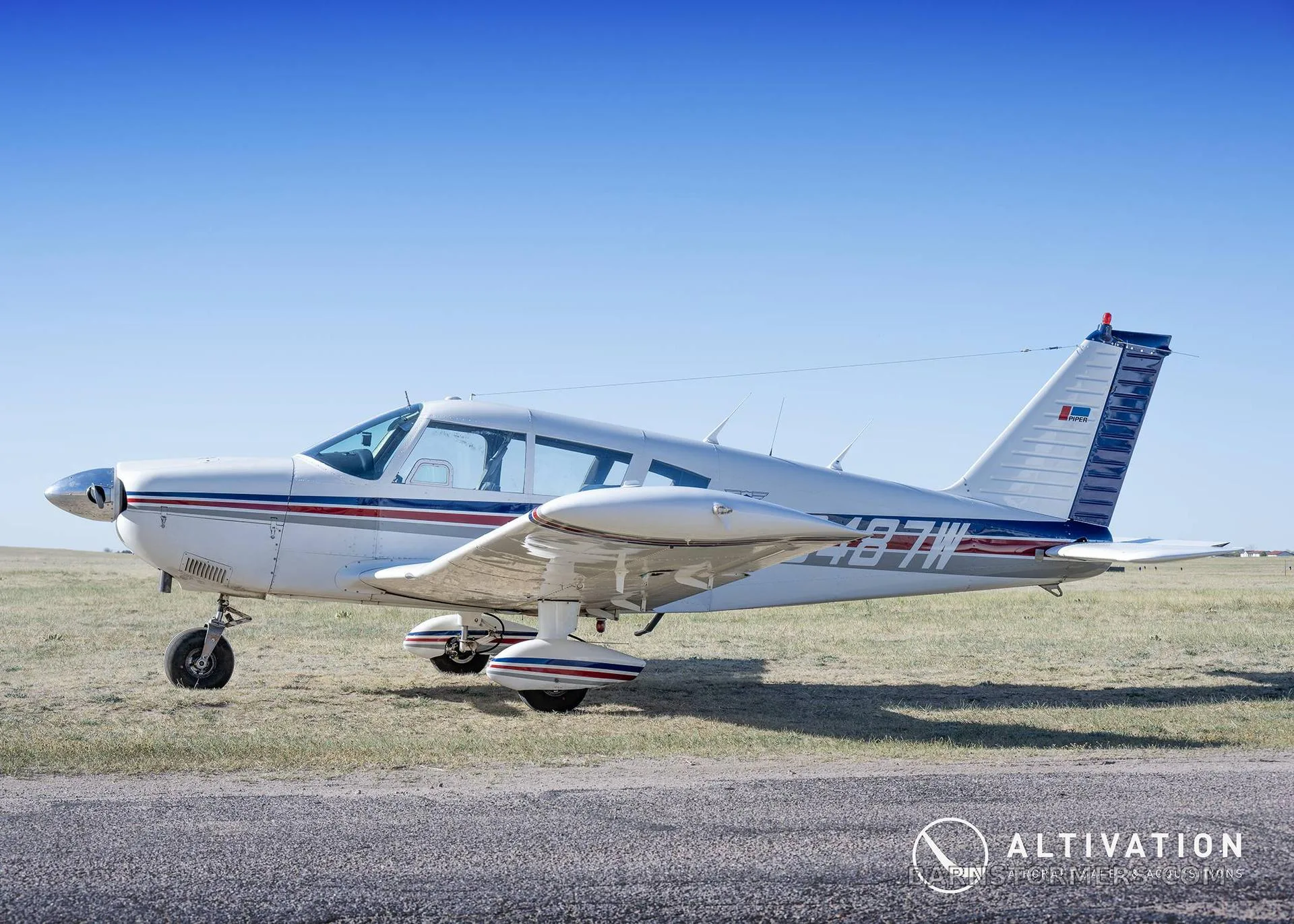 A white single-engine propeller aircraft with blue and red stripes is parked on a grassy area next to pavement under a clear blue sky.