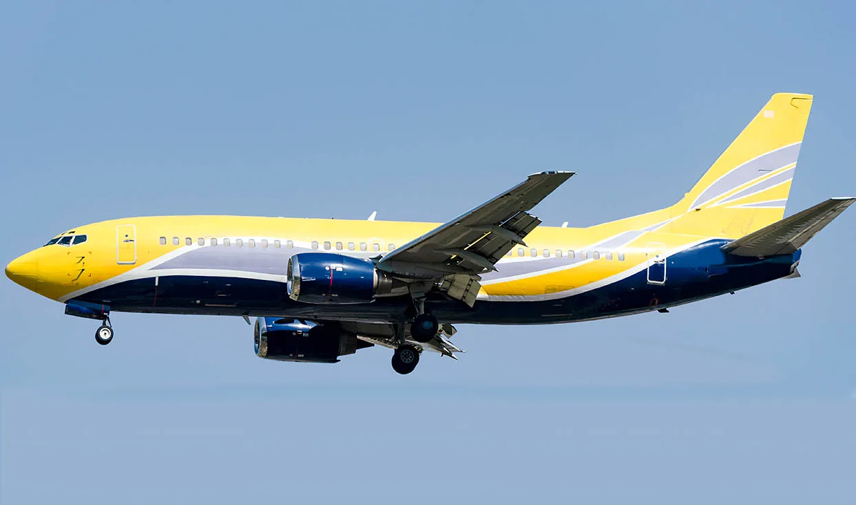 A Boeing 737 aircraft with a distinctive yellow, blue, and grey paint scheme flying with its landing gear extended against a clear blue sky.