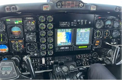 A close-up view of an aircraft's instrument panel, featuring numerous analog gauges and two digital displays, with a pilot's hand visible on the controls.