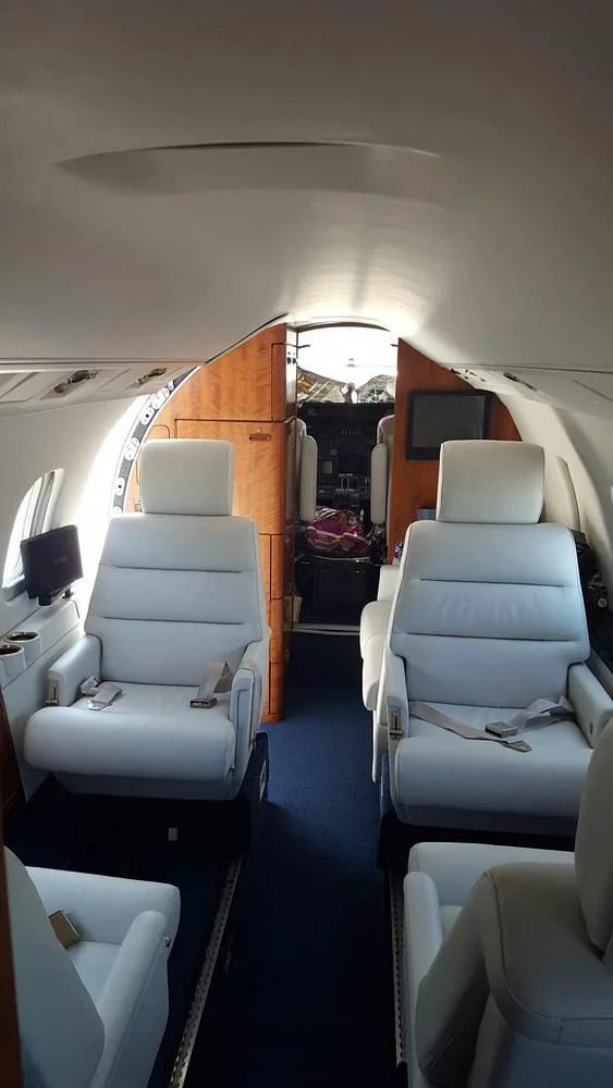 The interior cabin of a private jet with light grey leather seats and wood paneling, looking towards the cockpit.