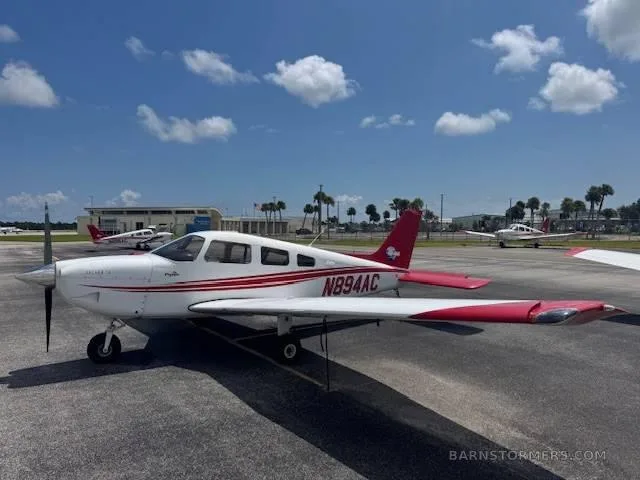 A white Piper Archer TX with red stripes and tail, registration N894AC, parked on an airport tarmac under a partly cloudy sky.