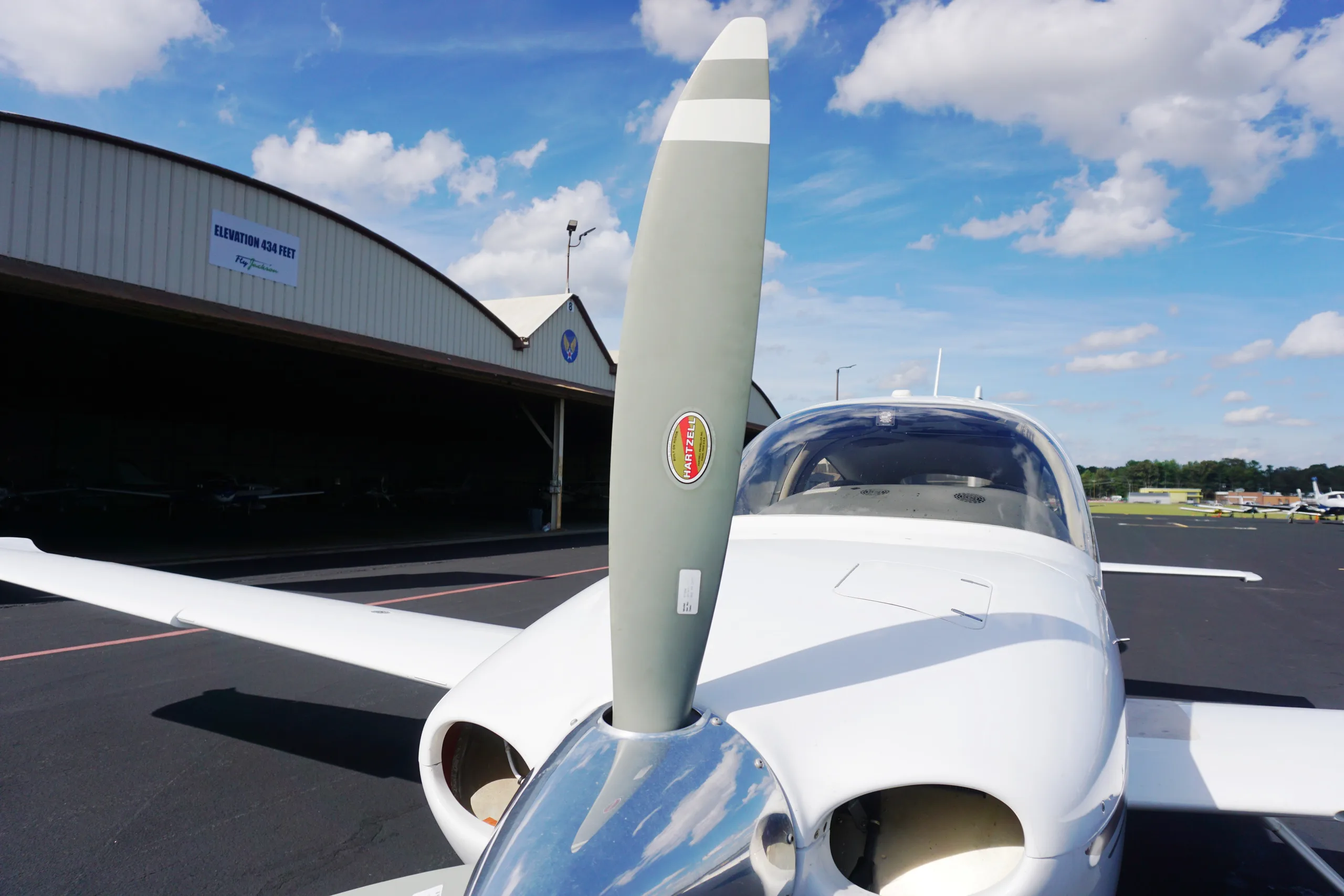 Front view of the propeller and nose of a white Cirrus SR22, parked on an airport tarmac in front of a hangar.