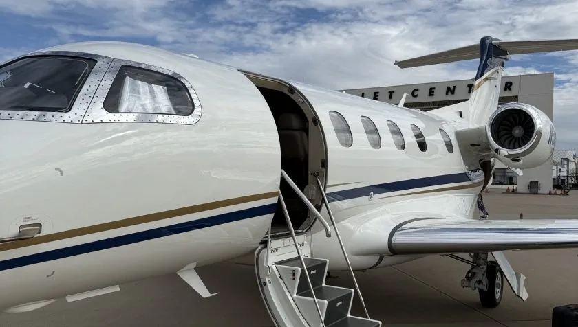 A close-up side view of a white Embraer Phenom 300 private jet with its passenger door open and stairs extended, parked at a jet center.