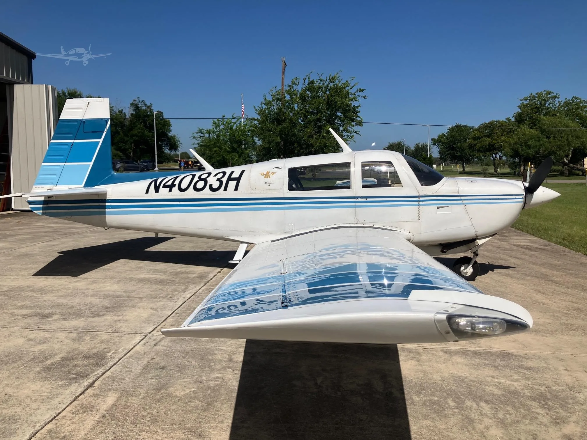 A white single-engine propeller aircraft with blue stripes and tail, registration N4083H, parked on a concrete apron next to a hangar.