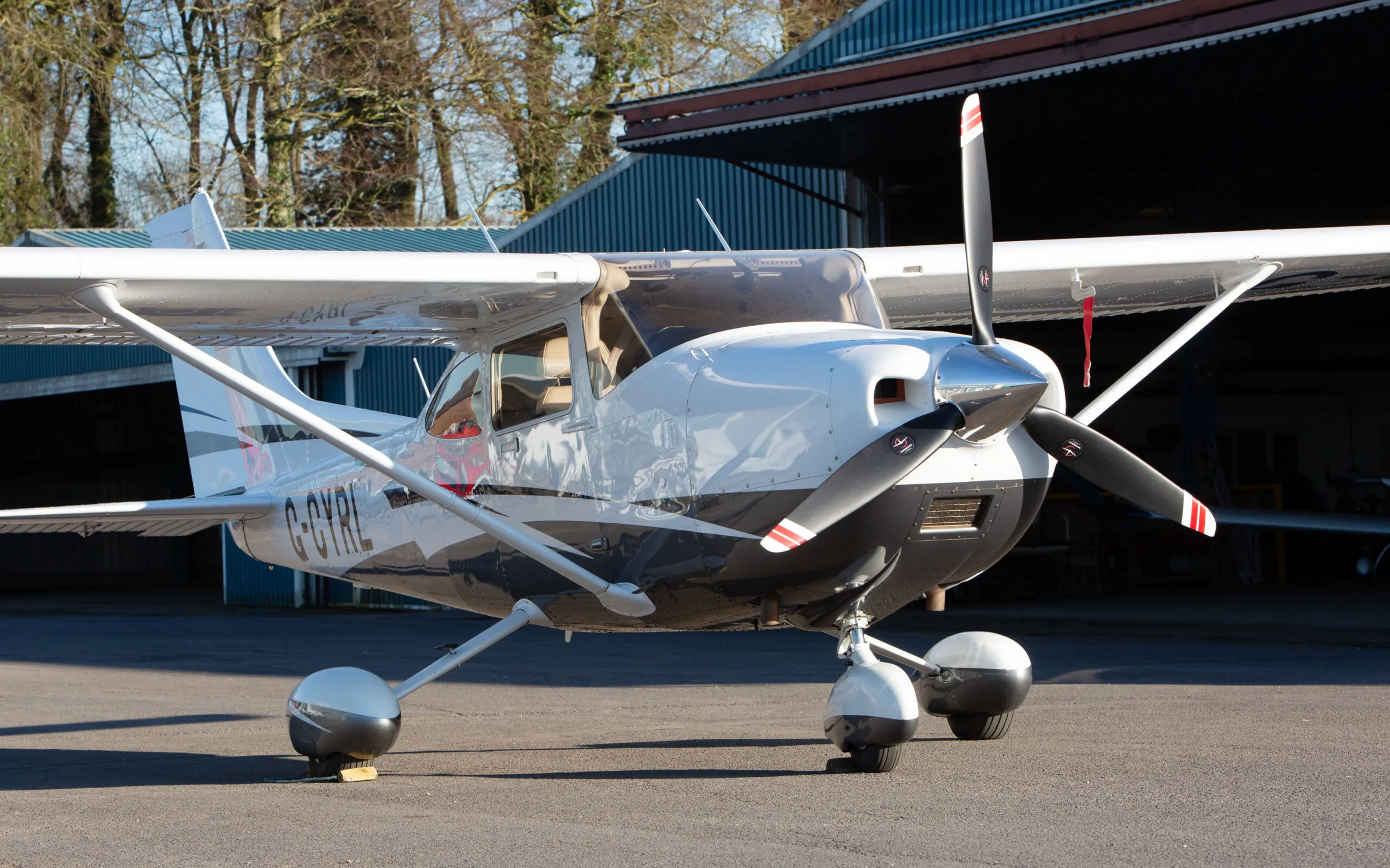 A white and black Cessna 182T (G-CYRL) parked on an airfield with trees in the background.
