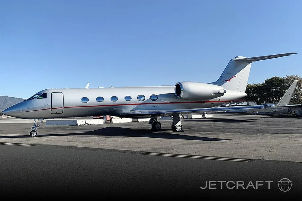 A silver Gulfstream G550 business jet with a red stripe parked on an tarmac under a clear blue sky.
