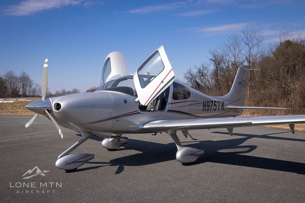 A silver Cirrus SR20 aircraft with registration N975TK parked on an asphalt runway with its doors open.