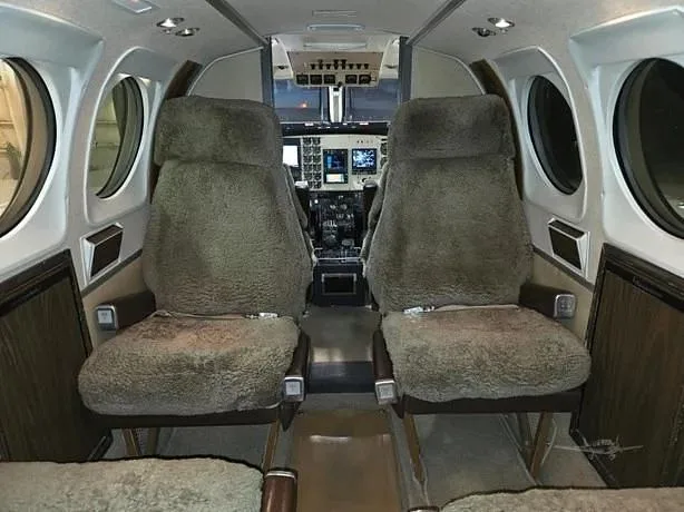 Two sheepskin-covered passenger seats are visible in the aircraft cabin, looking forward towards the cockpit.