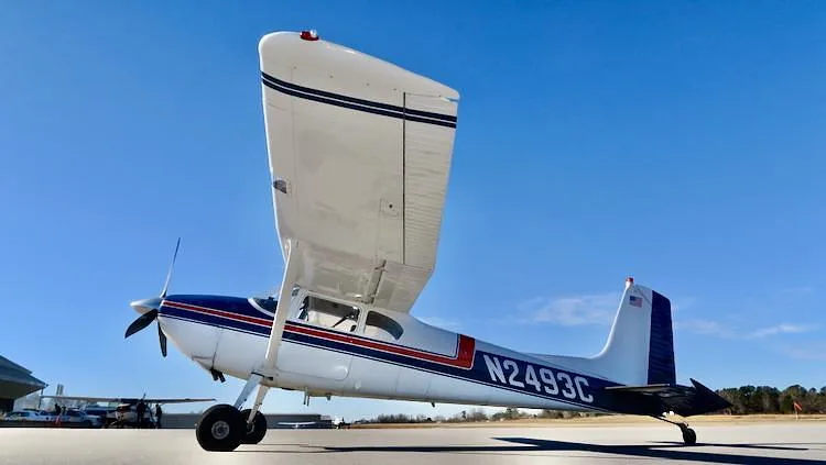 A low-angle, side view of a white and blue Cessna 180 Skywagon with red stripes, registration N2493C, parked on an airfield.