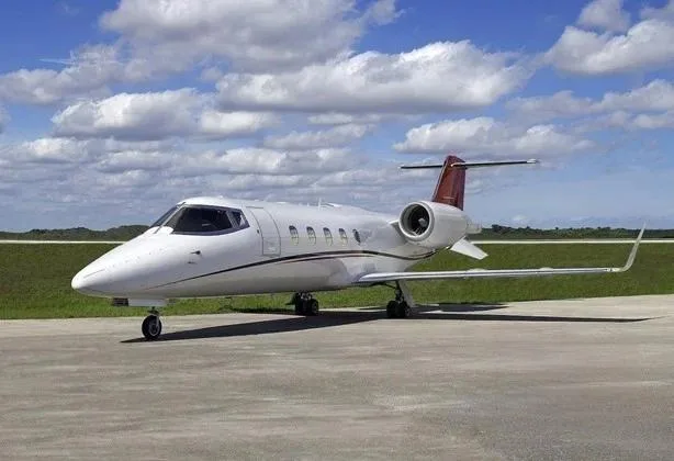 A white business jet with a red tail and dark stripe sits on an airport tarmac under a partly cloudy sky.