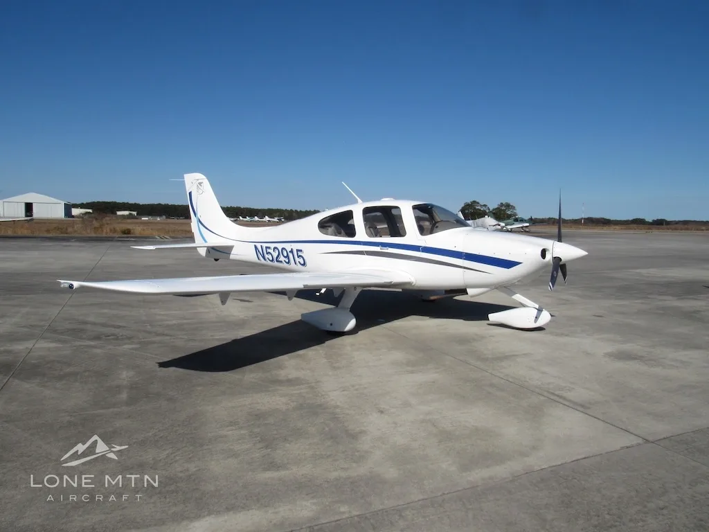 A white Cirrus SR20 aircraft with blue and grey stripes is parked on a concrete ramp under a clear blue sky.