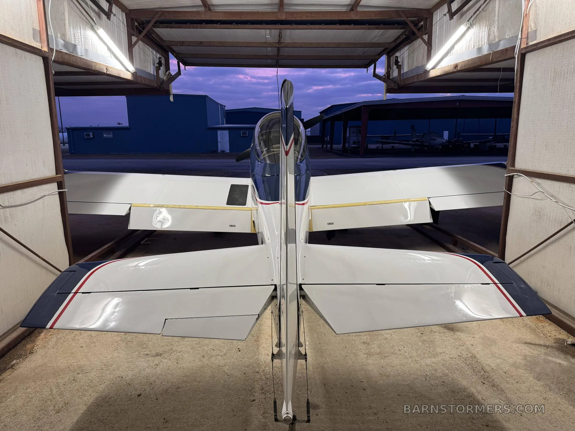 A blue and white single-engine aircraft viewed from the rear, parked inside a hangar with the hangar door open to a twilight sky.