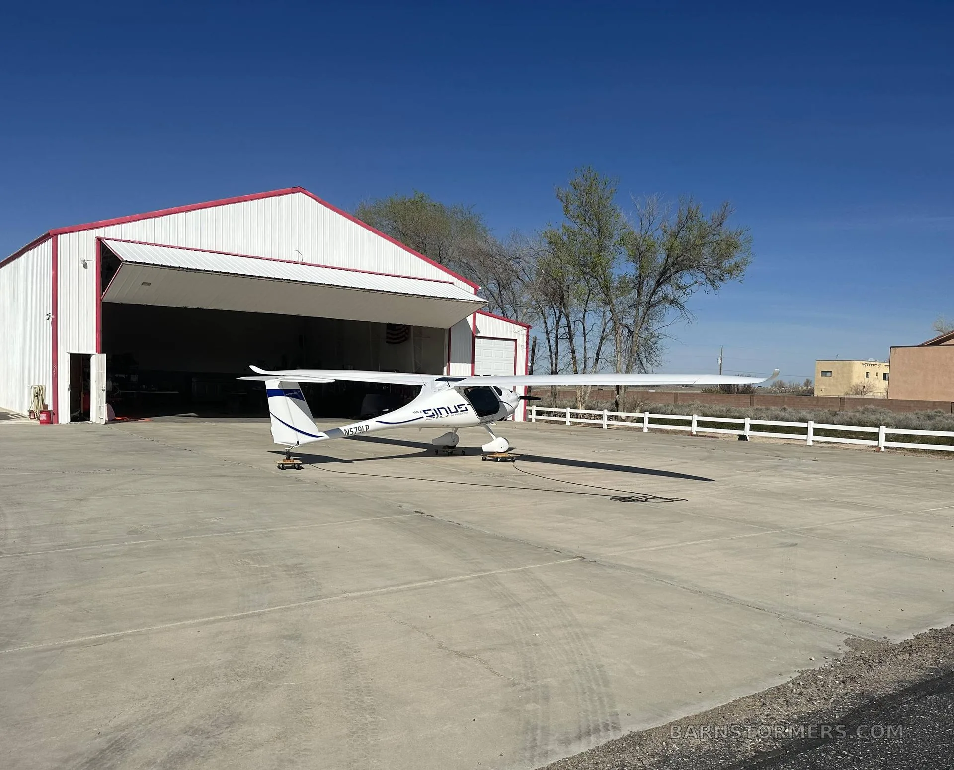 A white Pipistrel Sinus aircraft with blue accents is viewed from the rear-right, parked on an apron in front of a hangar.